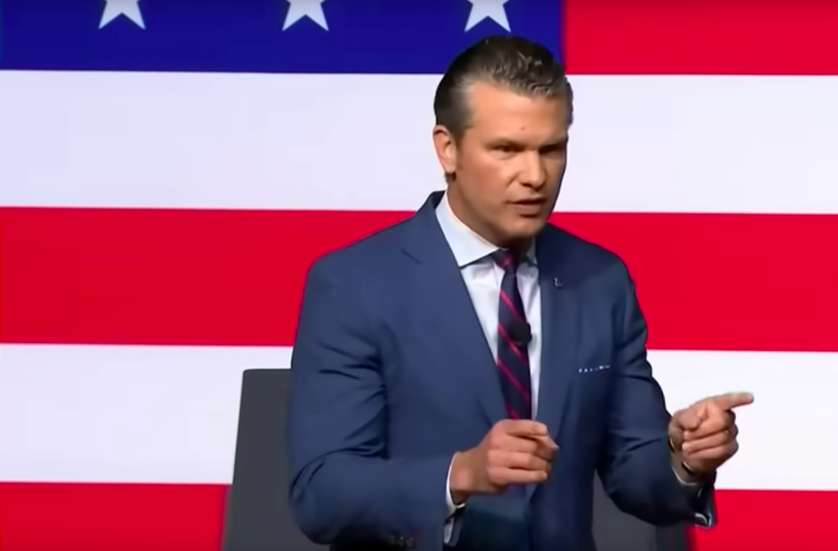 A tanned man with slicked back hair in an tight blue suit stands in front of a giant American flag giving a speech gesturing to the crowd