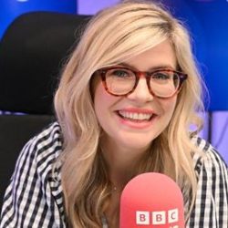 A woman with long hair and glasses smiling in front of a BBC microphone