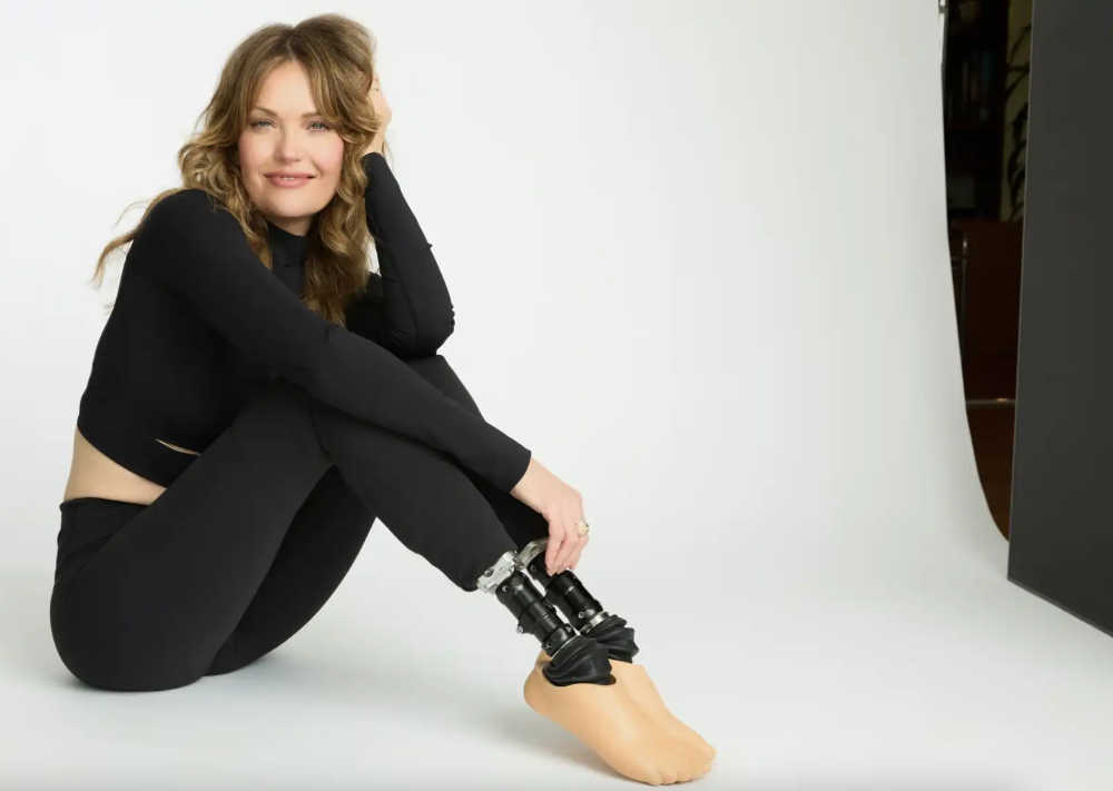 a woman with long wavy hair and prosthetic legs poses with her knees drawn up against a white photo backdrop