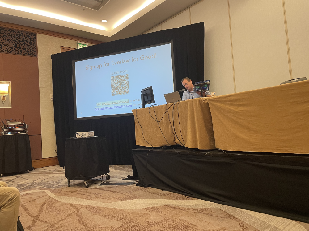 A man sits at a banquet table with a computer with a large screen next to him