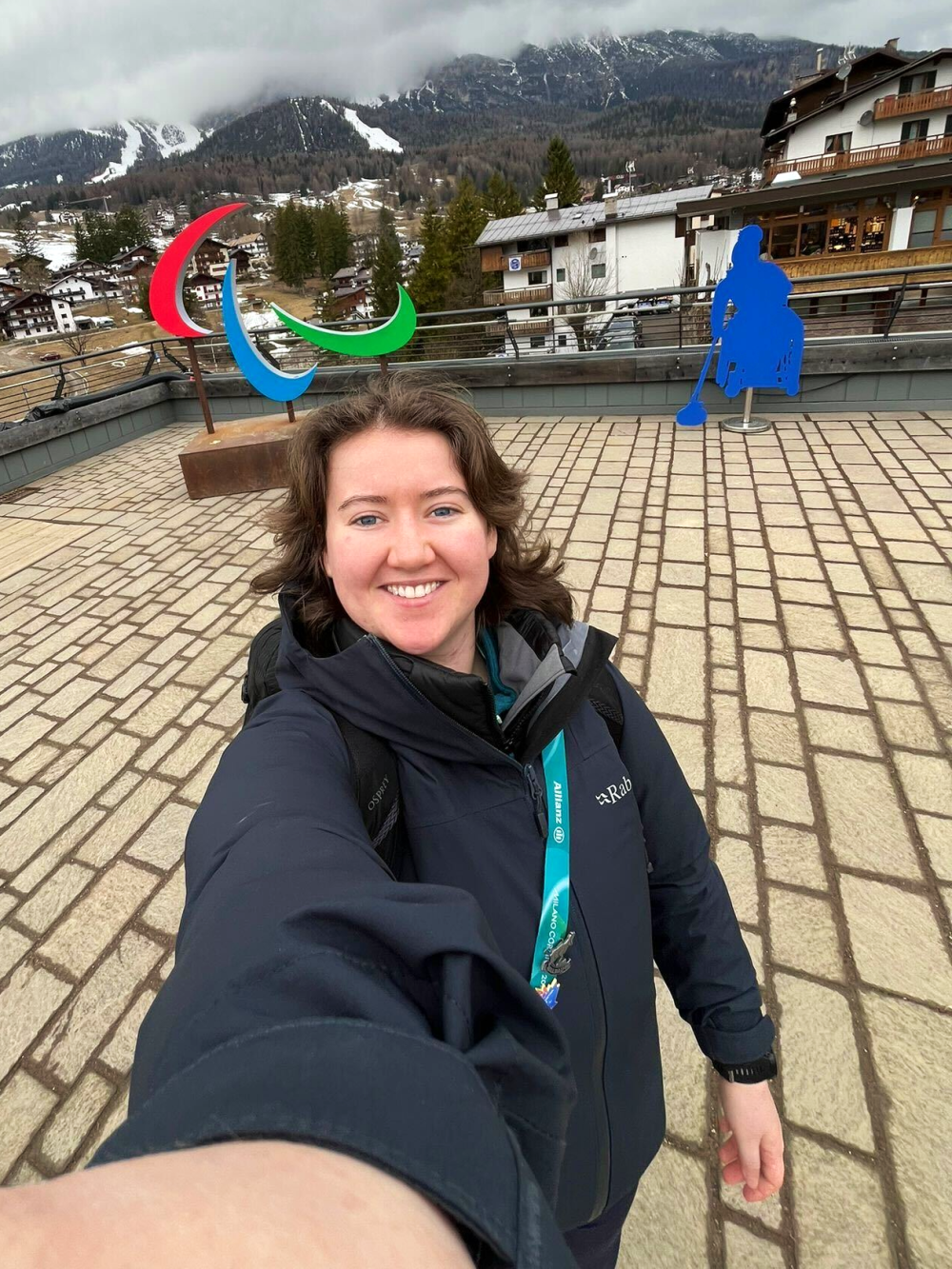 A reporter with shoulder-length curly chair taking a selfie in front of Paralympic Games sign