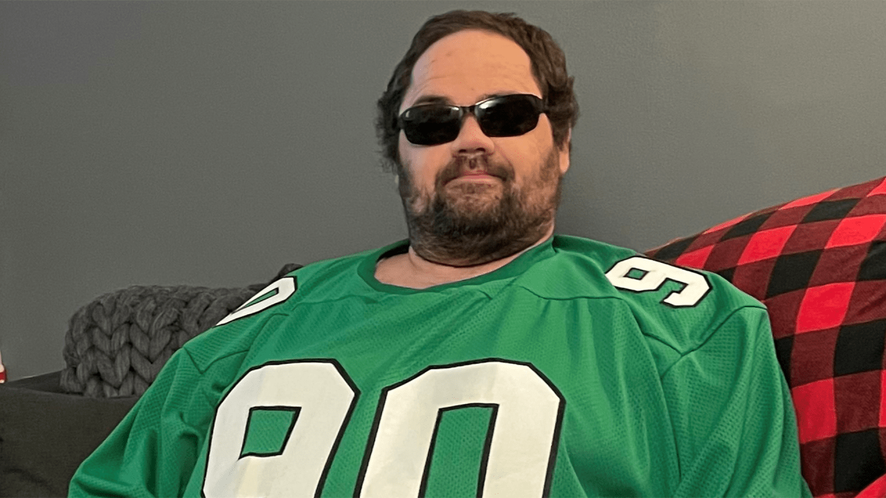 A blind man wearing a Philadelphia Eagles jersey and sunglasses sitting on a sofa