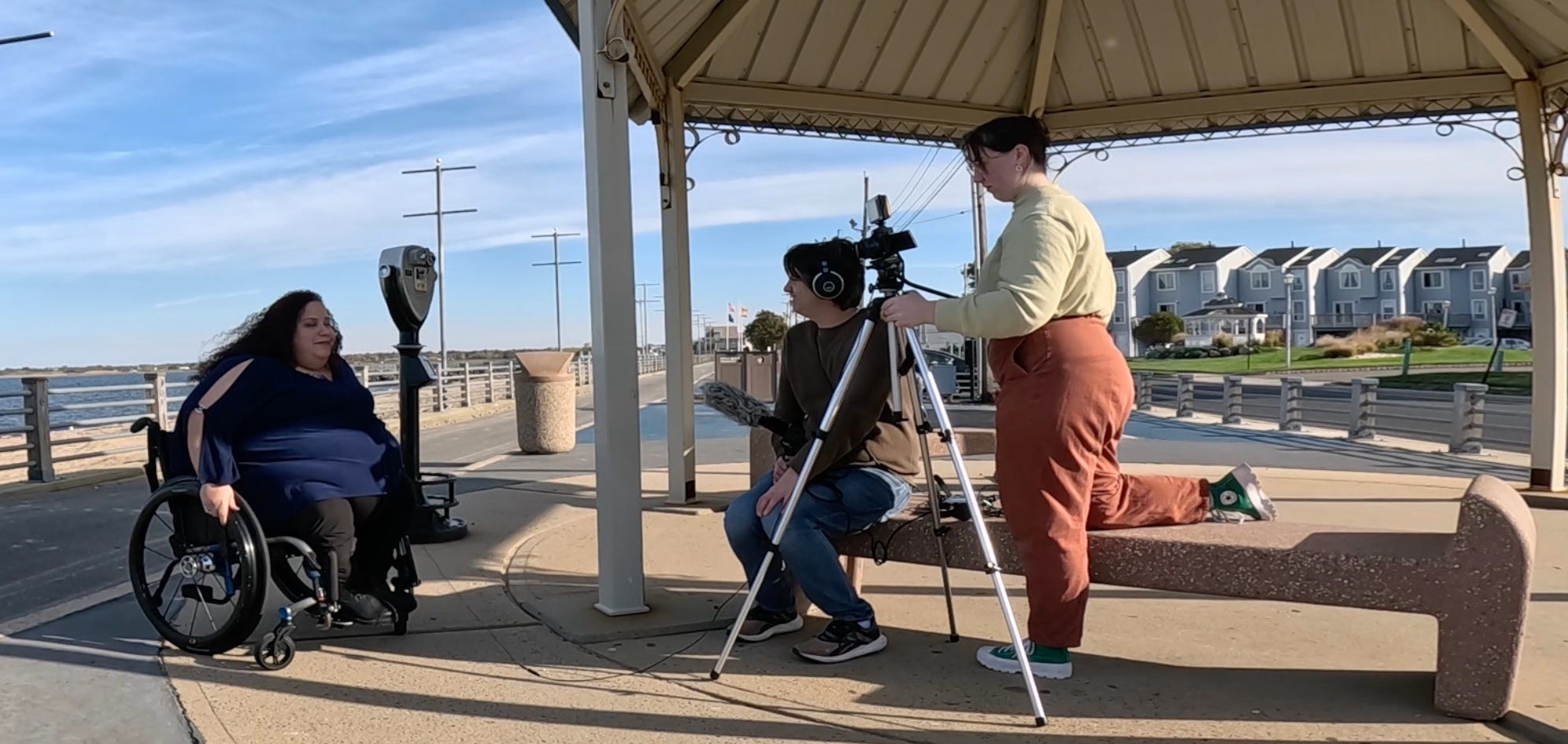 A woman sitting in a wheelchair on a board with the beach in the background is interviewed by a man in headphones holding a large microphone and a woman with a camera on a tripod