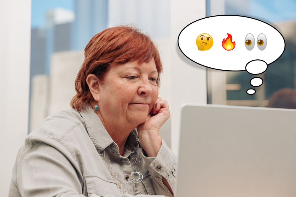 An older woman works on her laptop in an office with her red hair tucked behind her ear to reveal her hearing aid. A thought bubble appearing from the laptop shows three emoji: thinking face, fire, and eyes.
