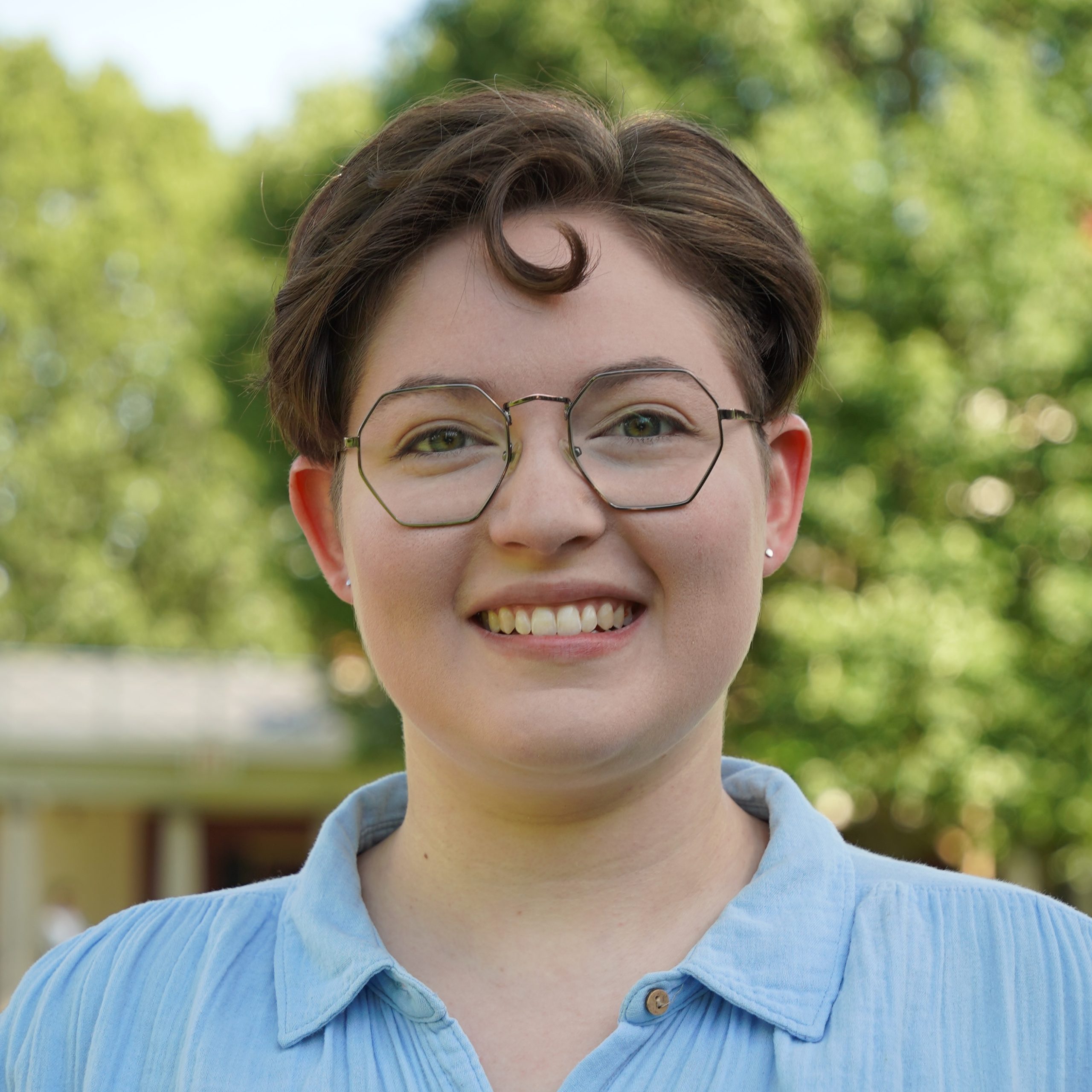 A person with short brown hair and glasses smiling with a curl looping over their forehead